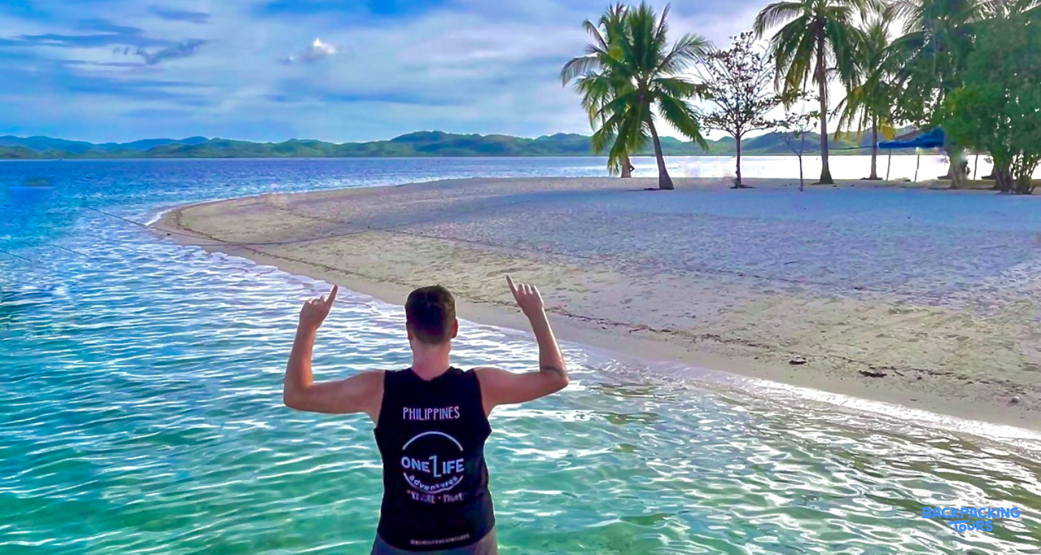 Backpacker in clear waters points to the stunning white sand beach and palm trees in Coron.