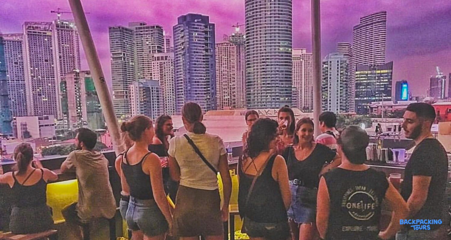 A group of backpackers socialize on a vibrant rooftop bar overlooking the Manila skyline at sunset.