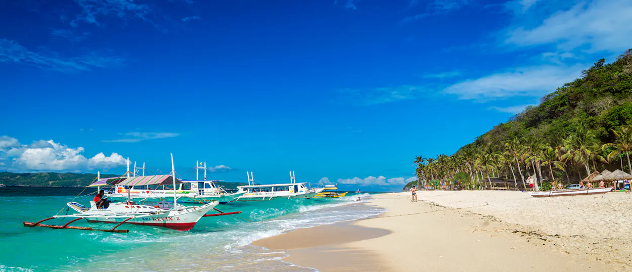 Traditional Philippine boats on turquoise water next to a white sand beach with palm trees.