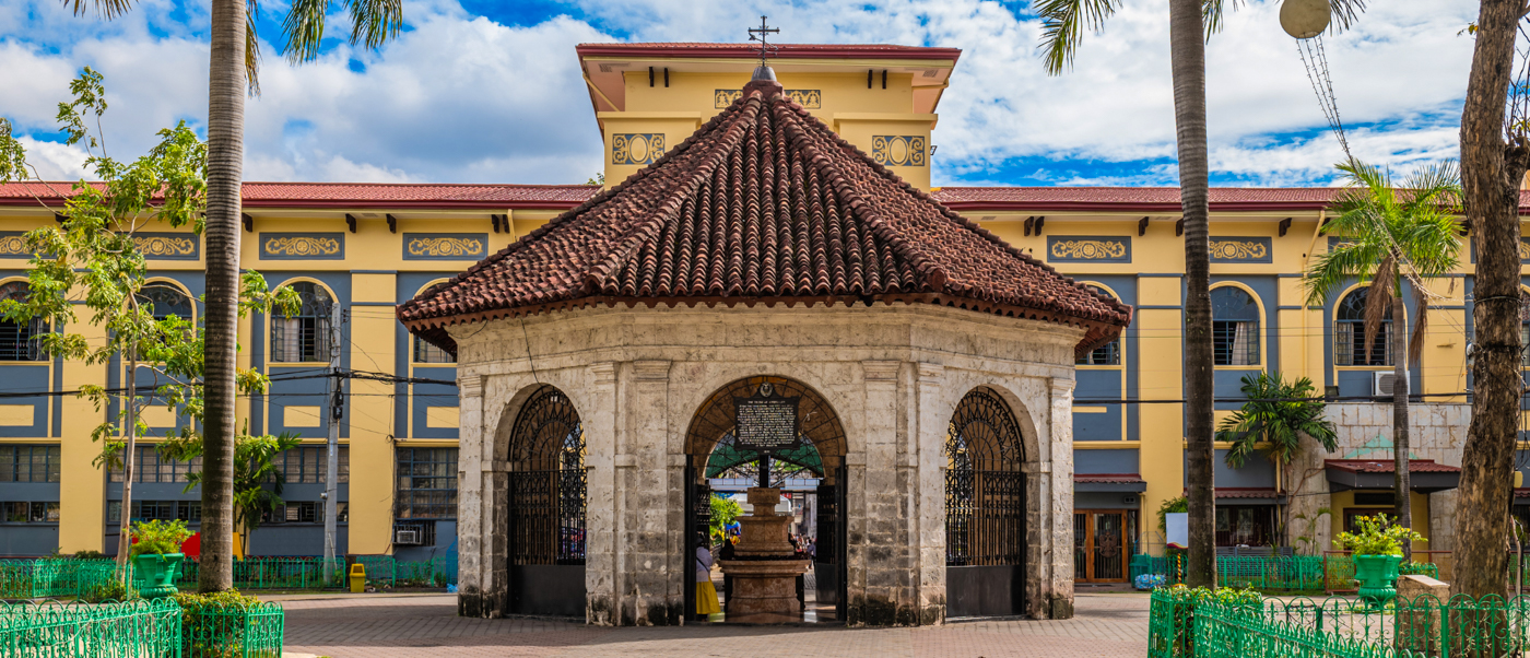 Historic Magellan's Cross shrine, an octagonal stone structure, stands before a yellow building in Cebu City.