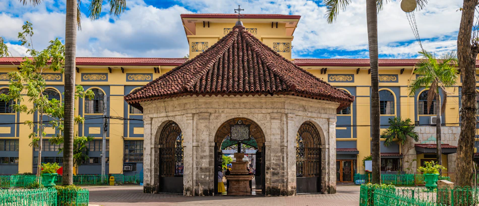 Historic Magellan's Cross shrine, an octagonal stone structure, stands before a yellow building in Cebu City.