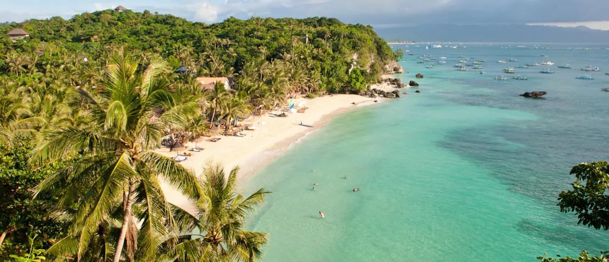 Palm trees frame a beautiful Boracay beach with white sand, clear turquoise water, and distant boats.