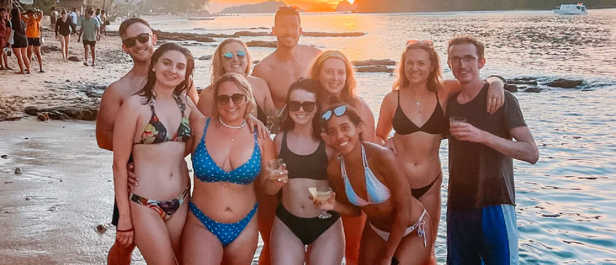 A diverse group of happy friends in swimwear posing on a beach at sunset, enjoying drinks.