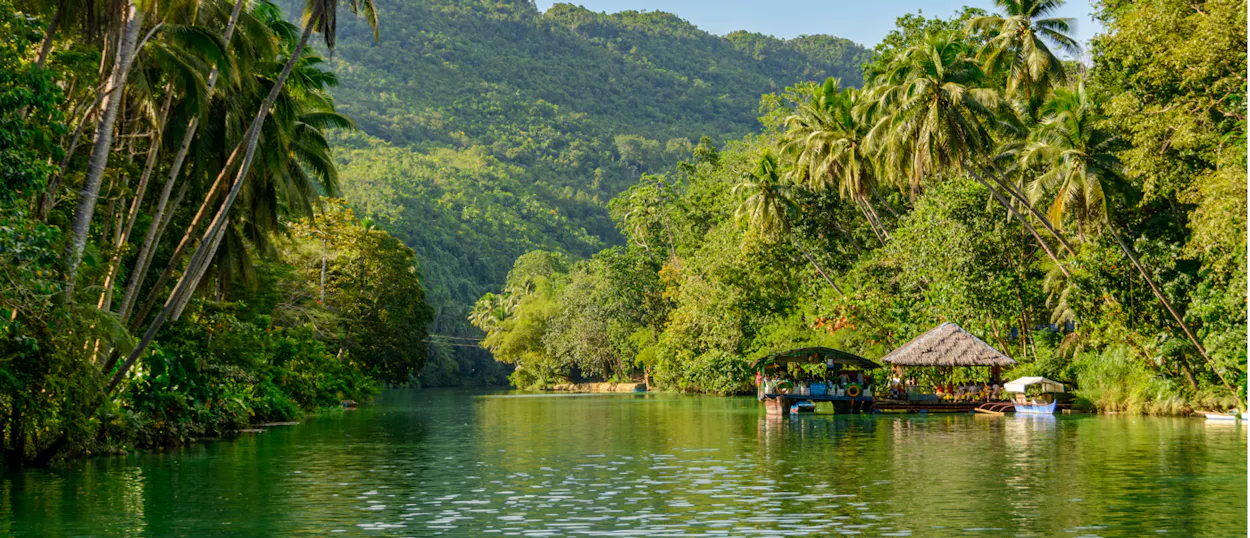A serene view of the Loboc River in Bohol, lined with lush tropical trees and floating huts.