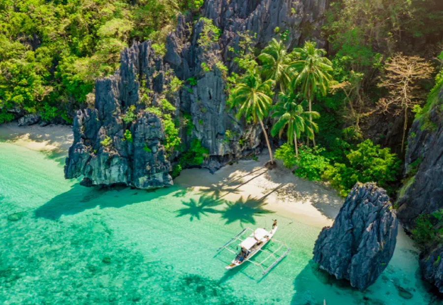 Tropical beach in the Philippines with turquoise water, limestone cliffs, palm trees, and a boat.