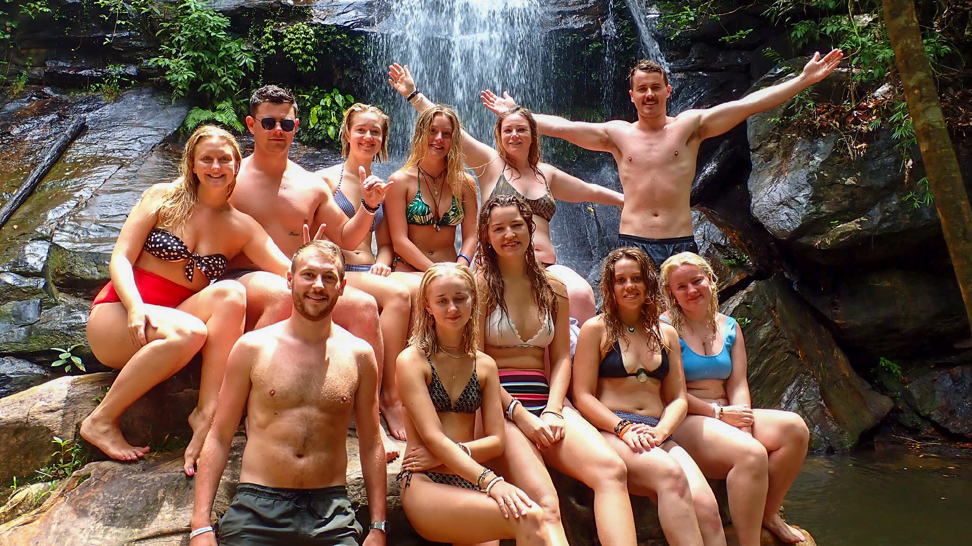A group of happy backpackers posing in swimwear in front of a beautiful waterfall in the Philippines.