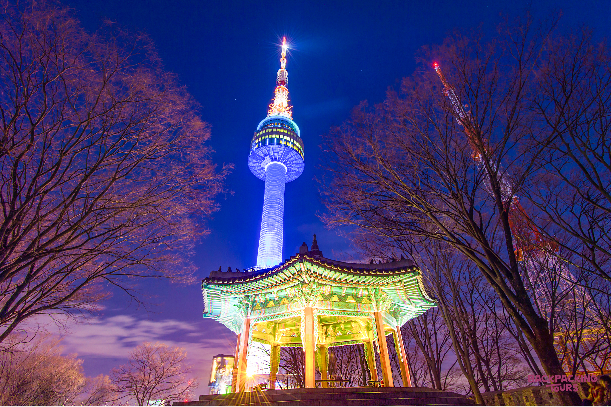 Namsan Tower glows blue at night, towering over a traditional pavilion and bare trees in Seoul.