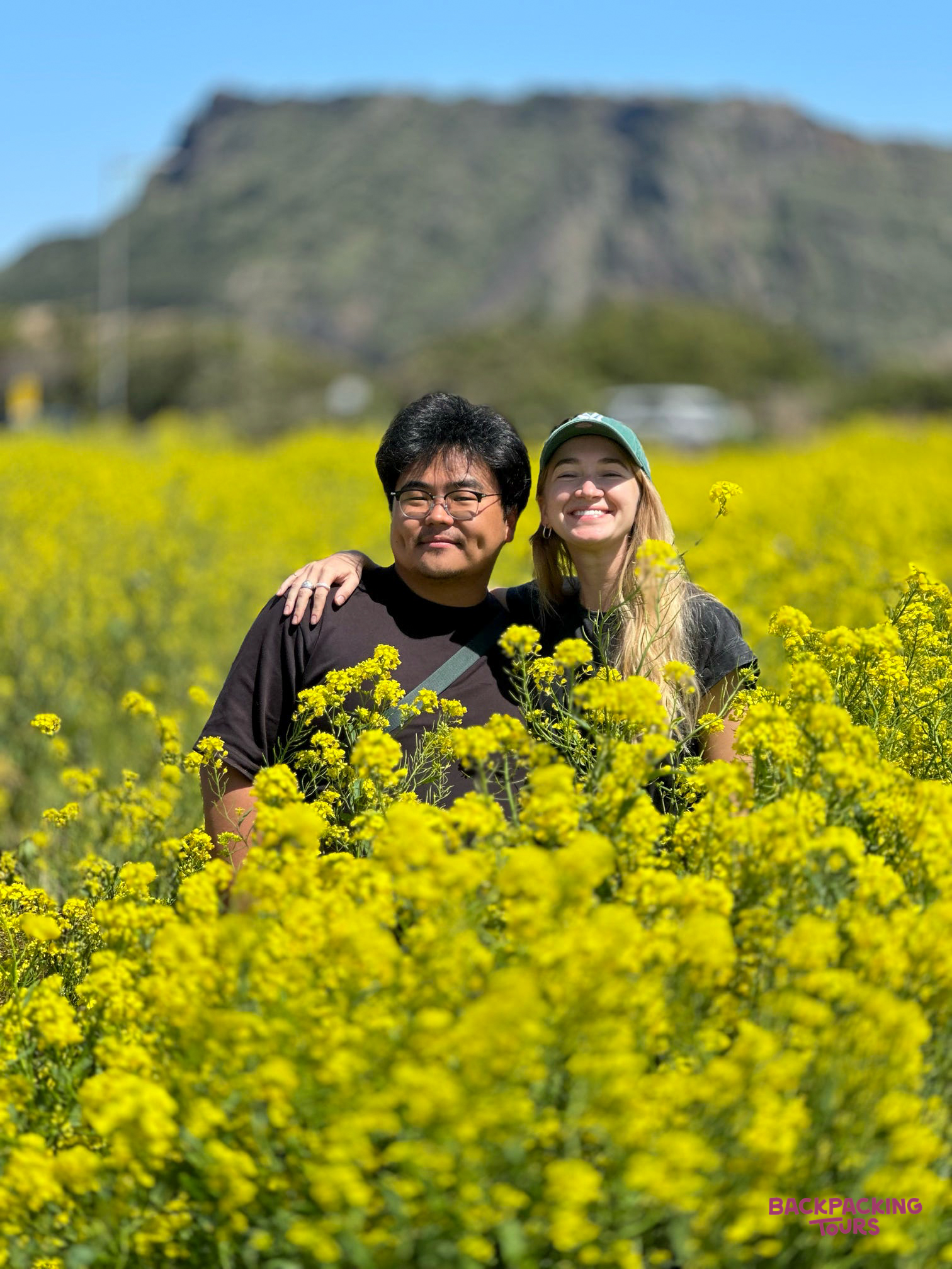 Two happy travelers pose in a vibrant yellow canola field on Jeju Island, with Seongsan Ilchulbong.