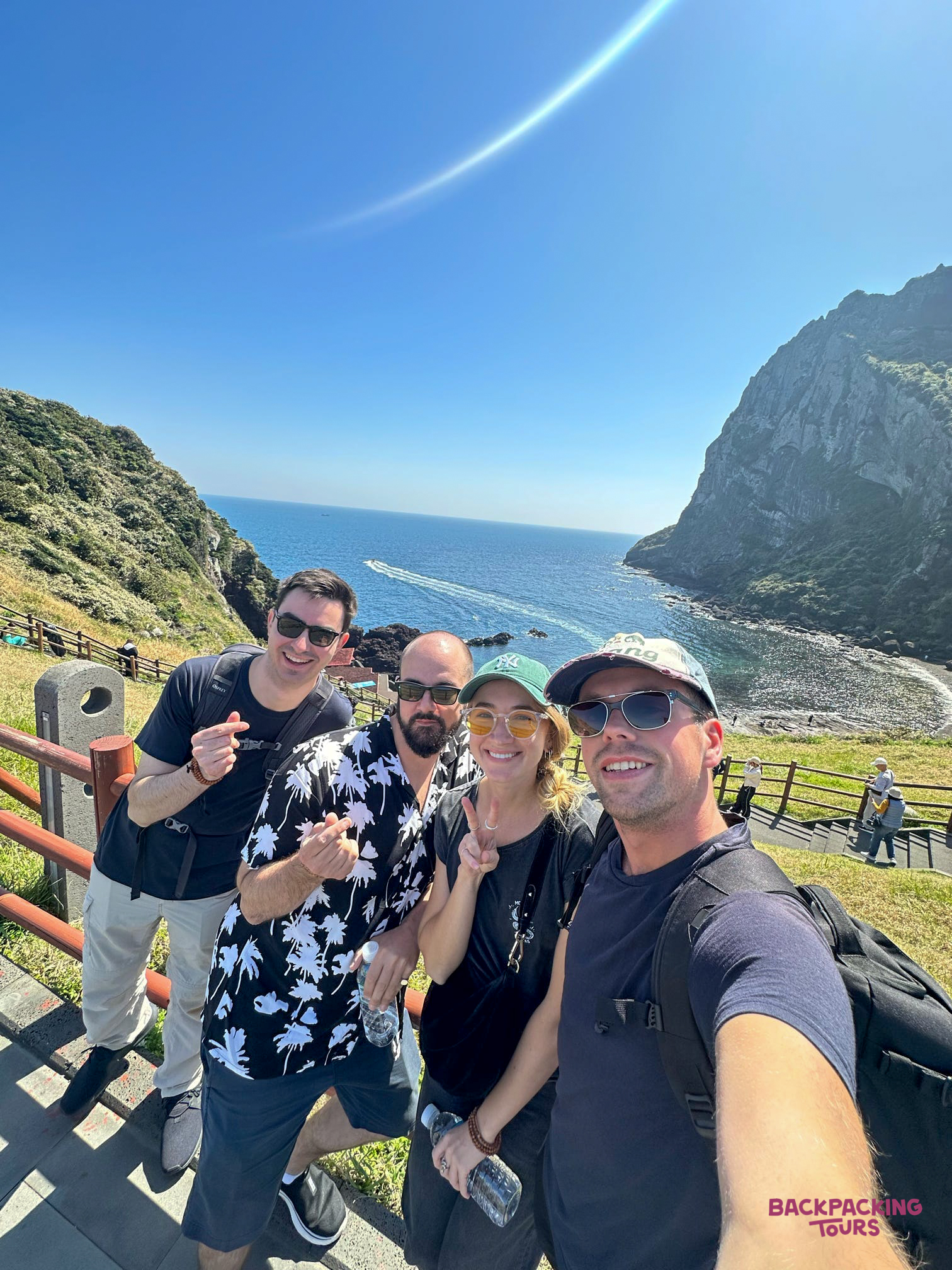 Backpackers enjoying a sunny day hike, posing for a selfie overlooking the ocean on Jeju Island.