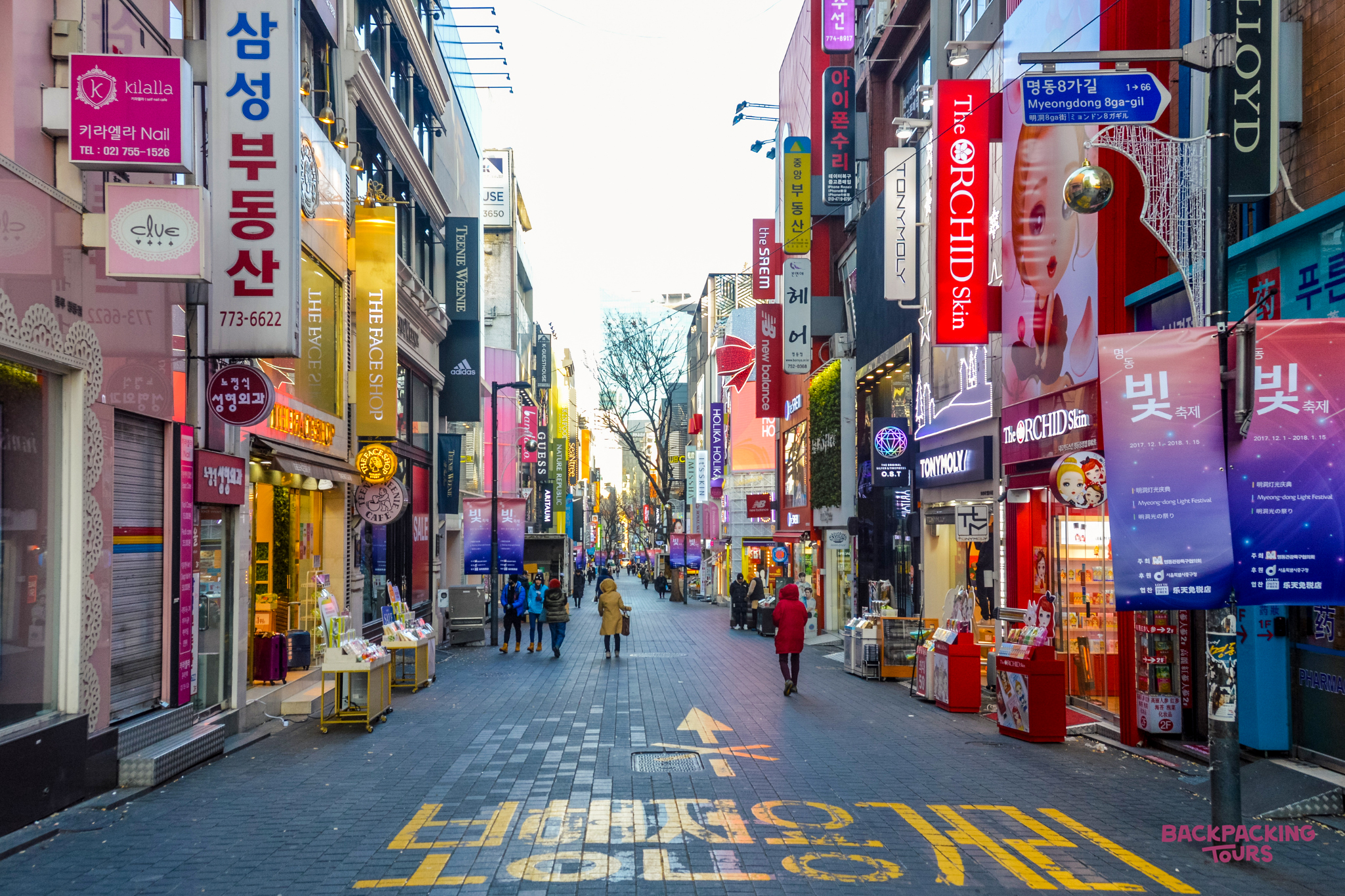 People stroll along a lively shopping street in Seoul, surrounded by bright signs and shops.