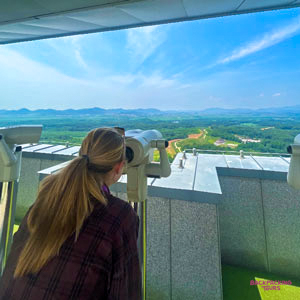 A backpacker peers through a telescope, observing the vast, green landscape of the Demilitarised Zone.