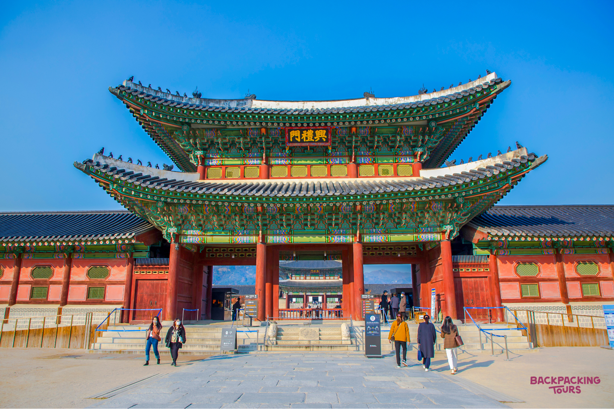 Tourists approach a grand, traditional Korean palace gate with vibrant colors and intricate architecture.