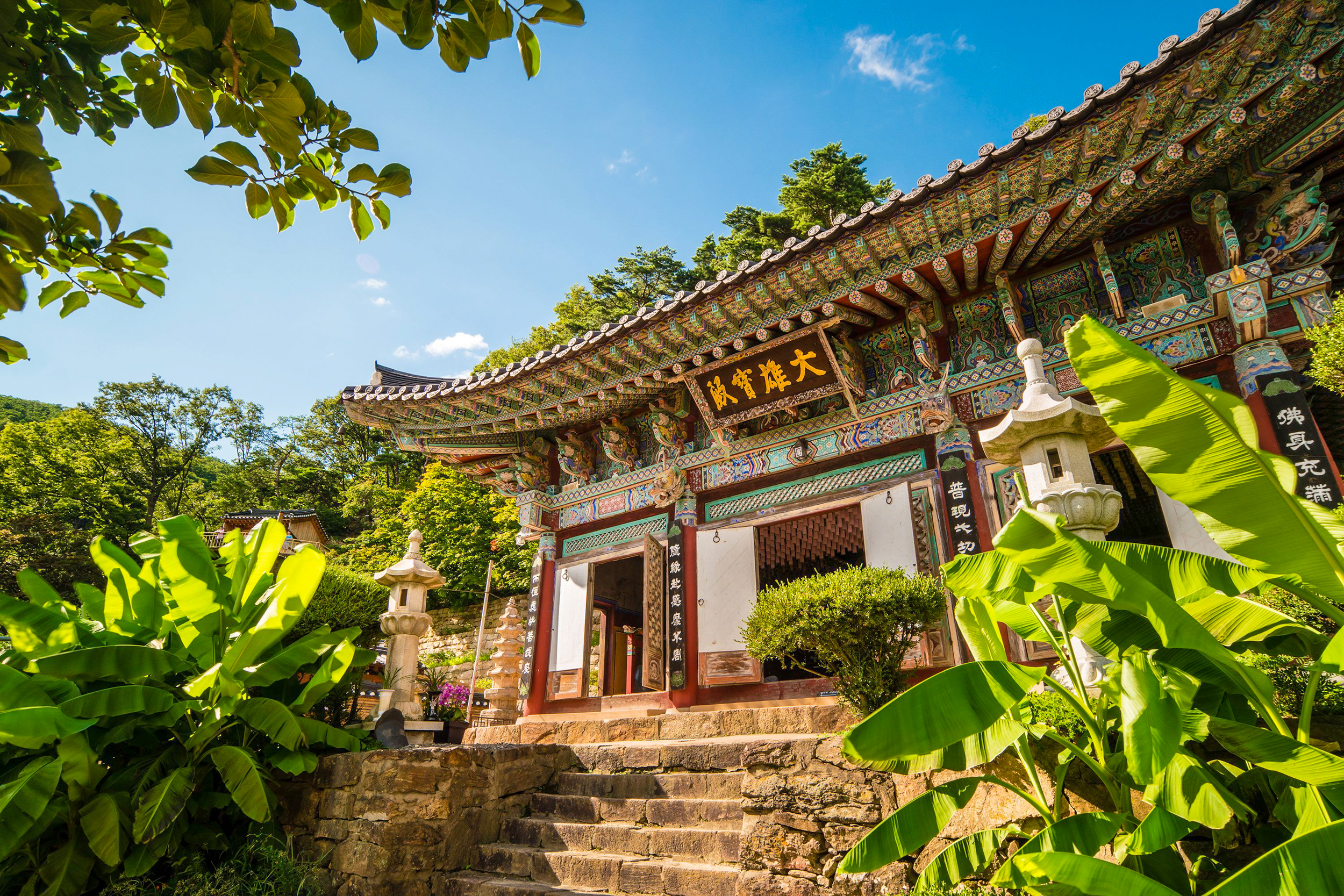 Ornate Geumsunsa Temple building nestled among vibrant green foliage and stone lanterns on a sunny day.