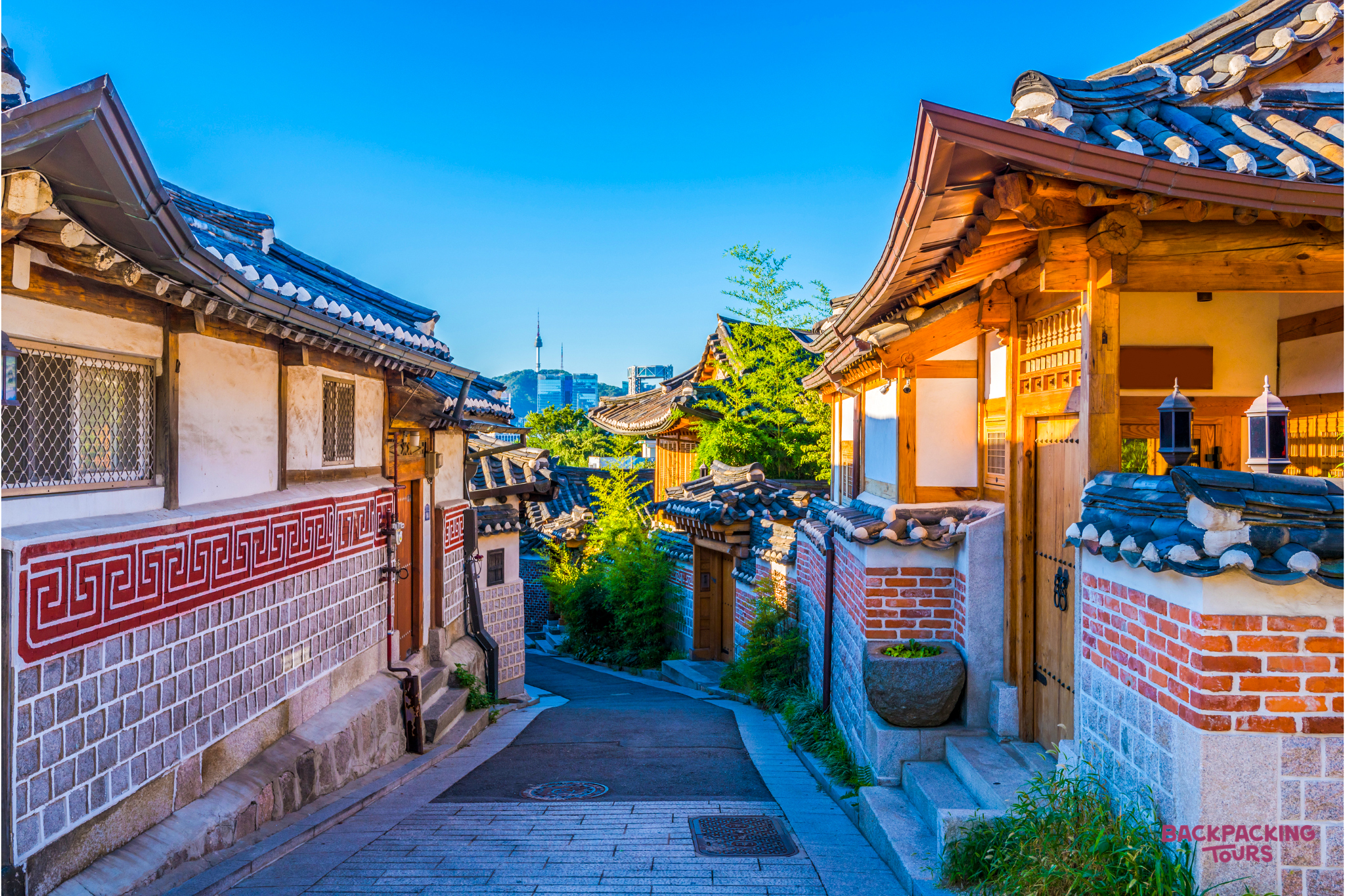 Traditional Korean Hanok houses line a narrow street, their tiled roofs contrasting with a distant modern city tower.