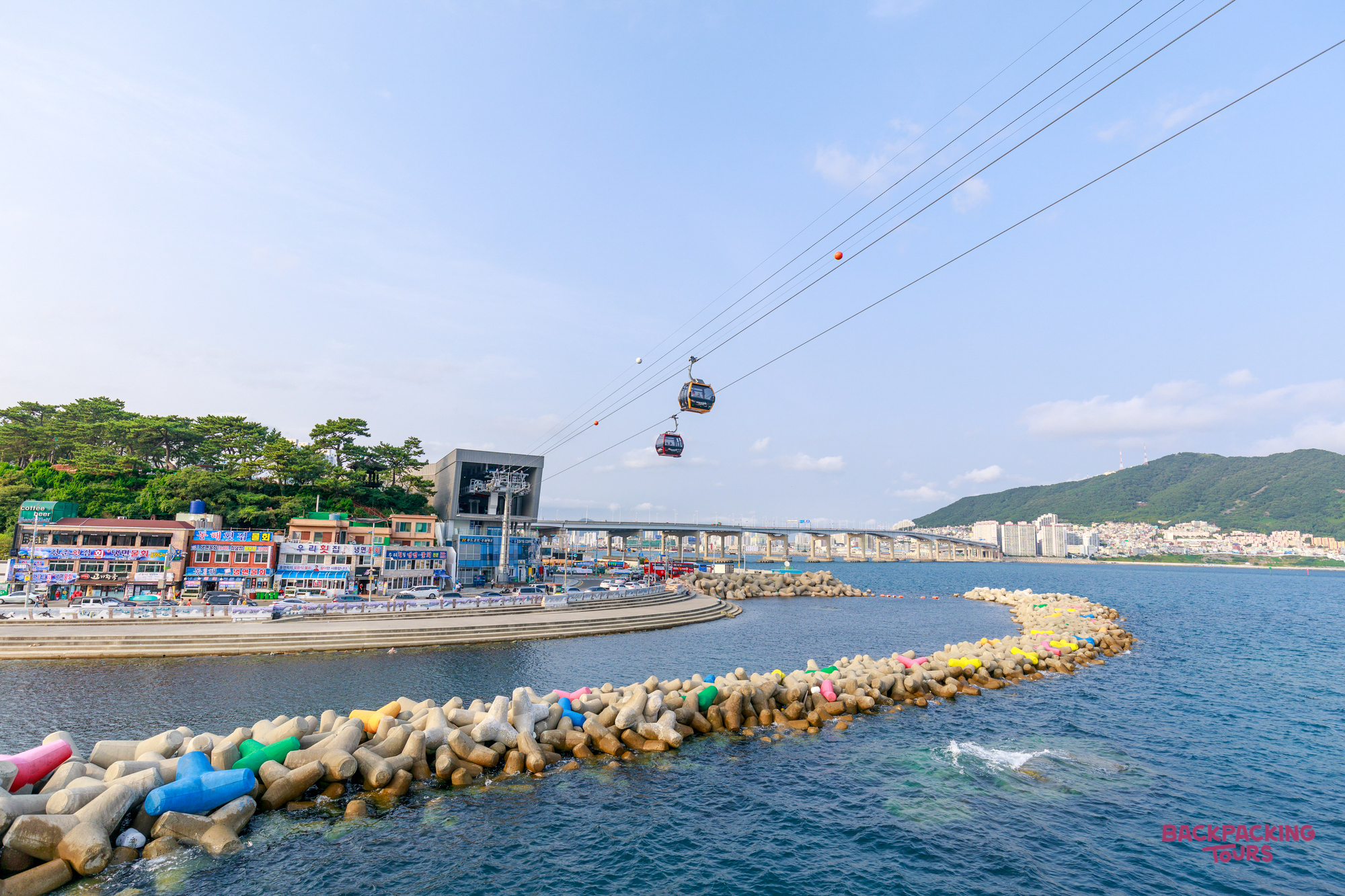 Cable cars glide over the vibrant blue waters of Busan, with colorful tetrapods lining the coast.