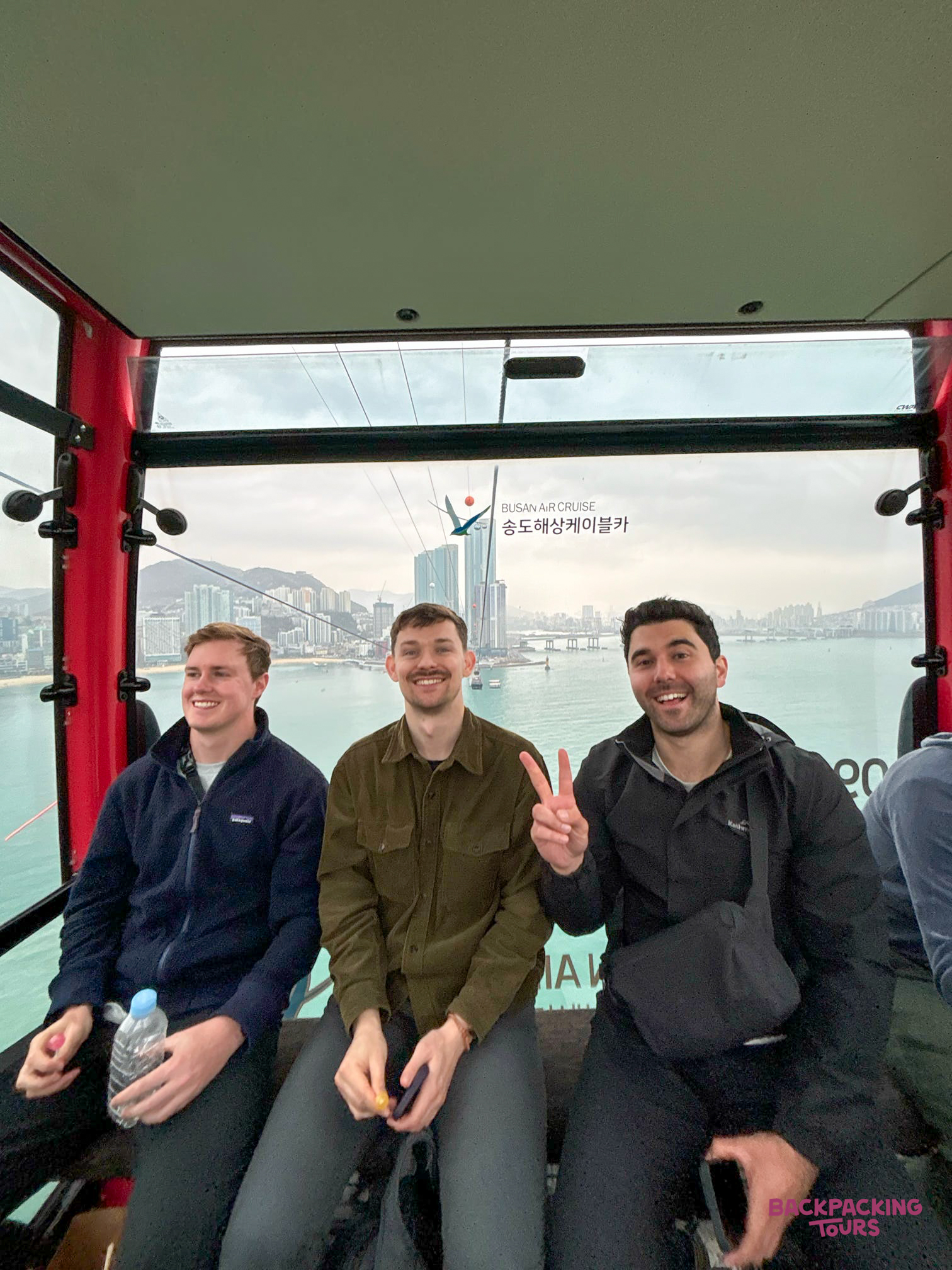 Three happy men riding the Songdo cable car, enjoying views of Busan city and the ocean.