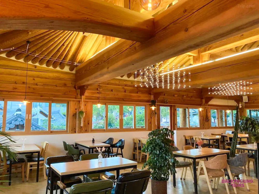 Warm wooden interior of a traditional Hanok guesthouse in Jeonju, with tables, chairs, and large windows.