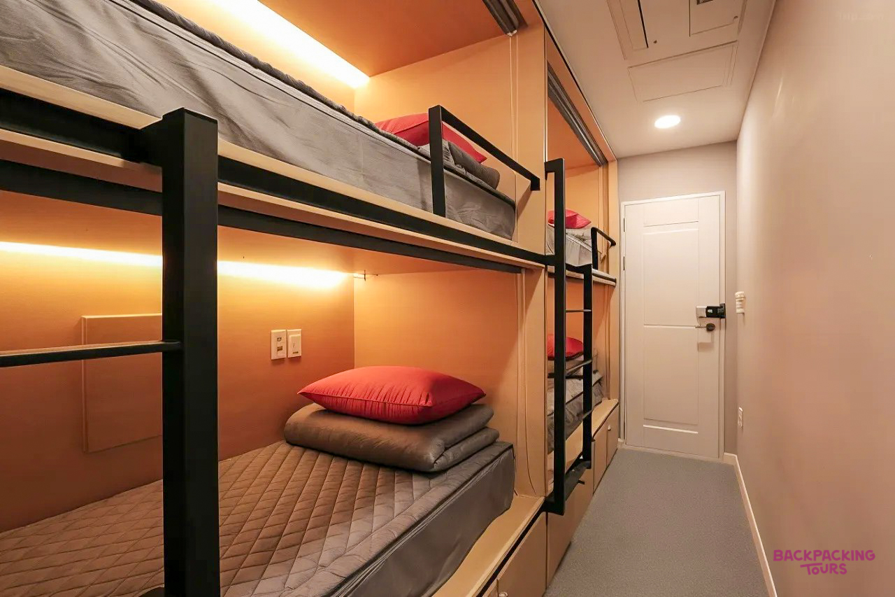 A modern hostel dormitory room with bunk beds, each featuring a red pillow and individual lighting.