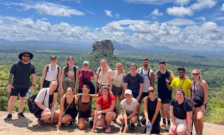 A large group of happy hikers posing on Pidurangala Rock with Sigiriya in the background.