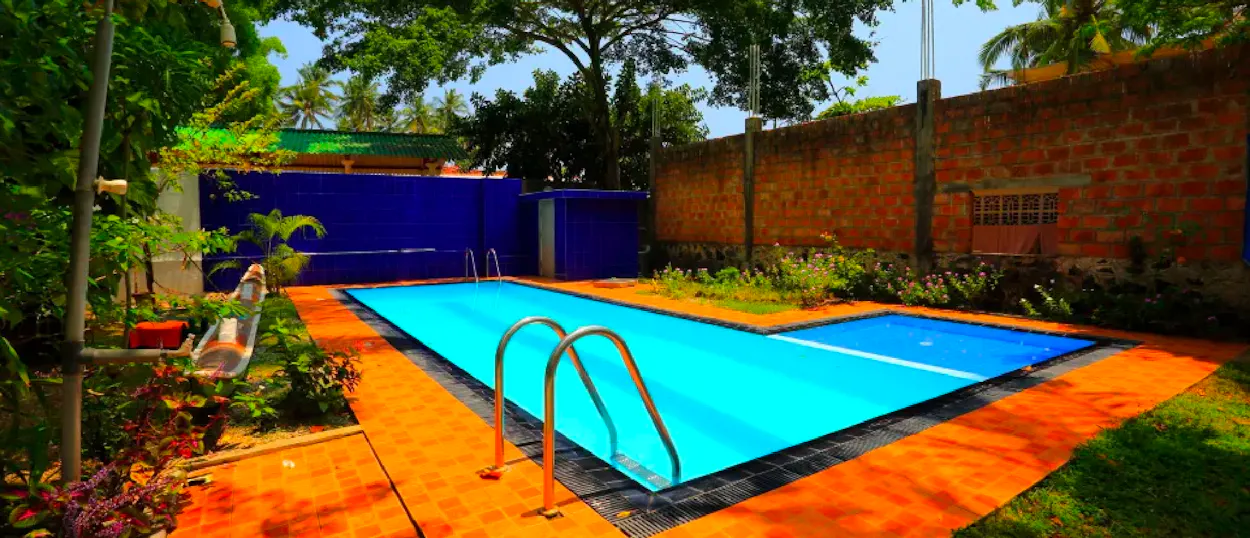 A vibrant blue swimming pool with orange paving surrounded by lush green tropical plants.