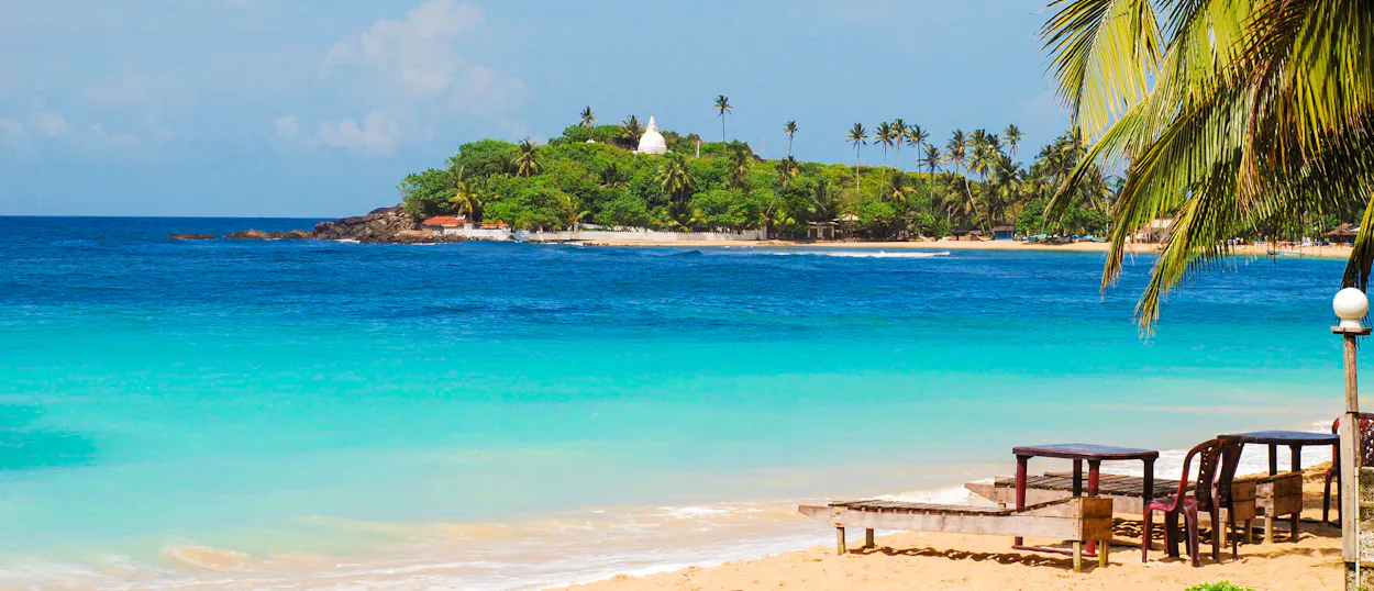 Golden sand beach in Weligama, Sri Lanka, with clear turquoise water and a lush green island.