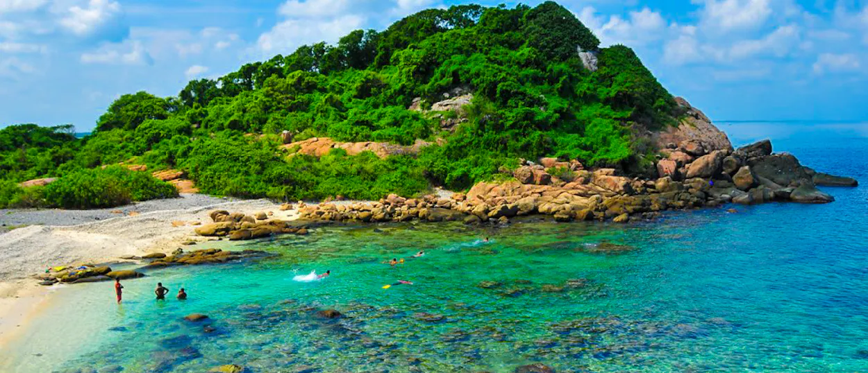 People swim in the clear turquoise waters of Weligama beach with a lush green island.
