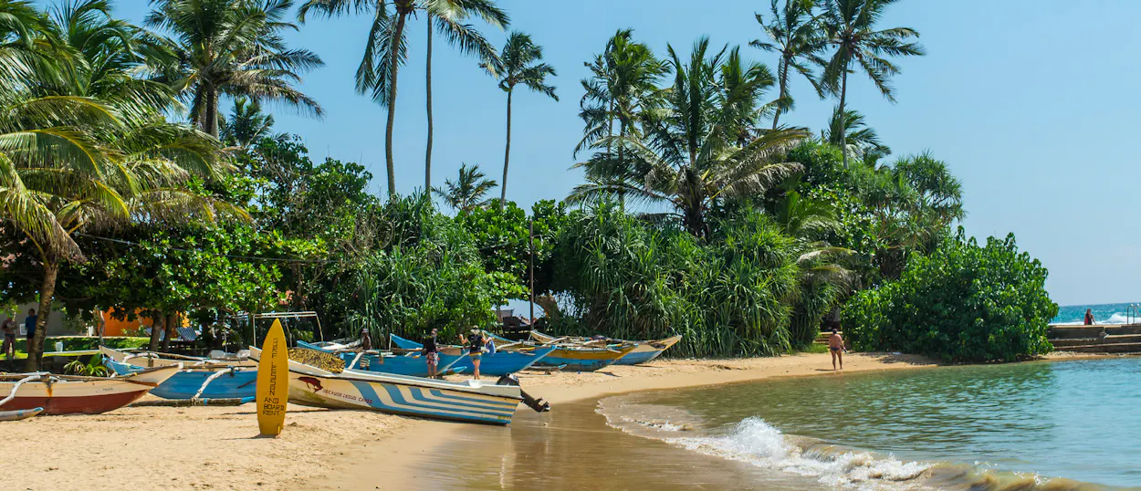 Traditional fishing boats and a yellow surfboard on a sunny tropical beach with palm trees in Unawatuna.