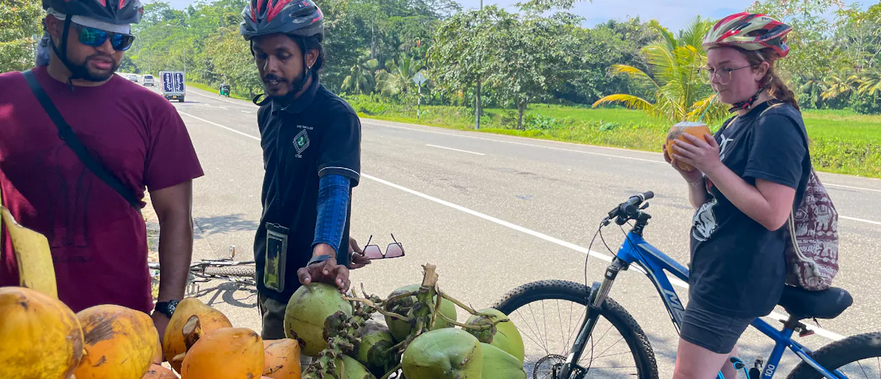 Two cyclists, wearing helmets, enjoy refreshing coconuts from a roadside stall in Unawatuna, Sri Lanka.