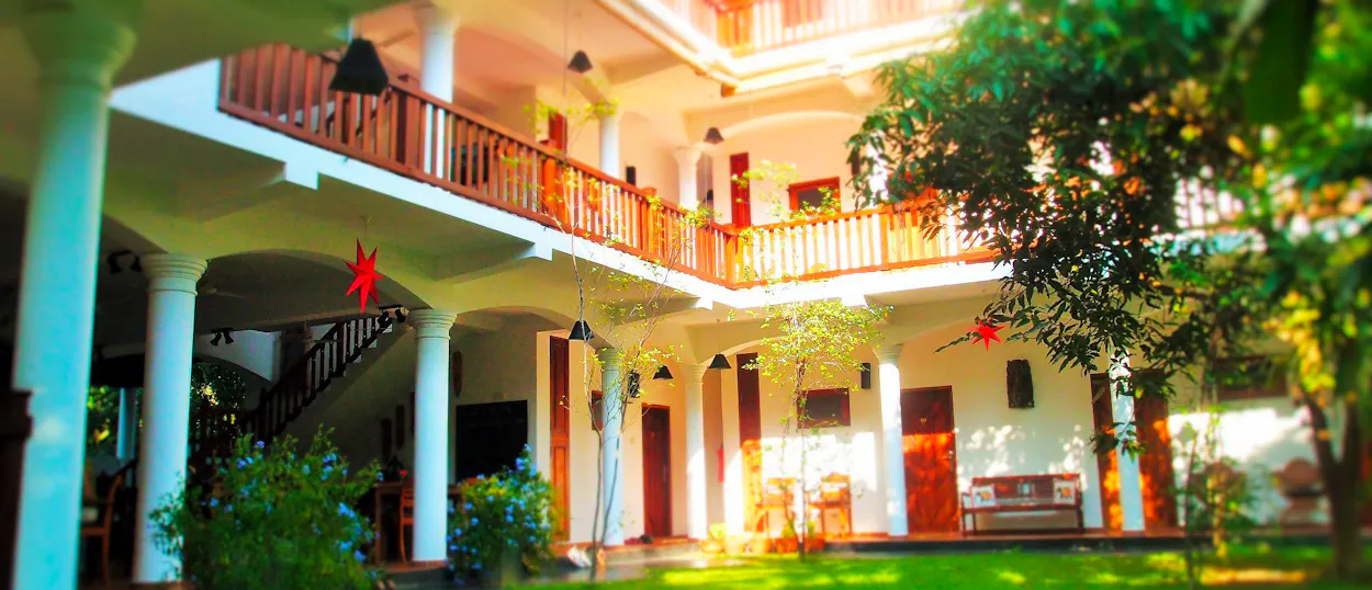 A beautiful white multi-story hotel with wooden balconies surrounds a lush green courtyard in Unawatuna.