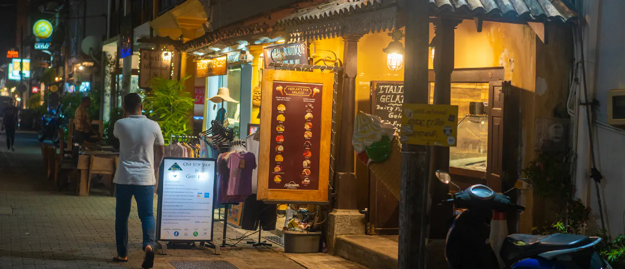 Man walks past brightly lit shops on a charming street in Galle at night.