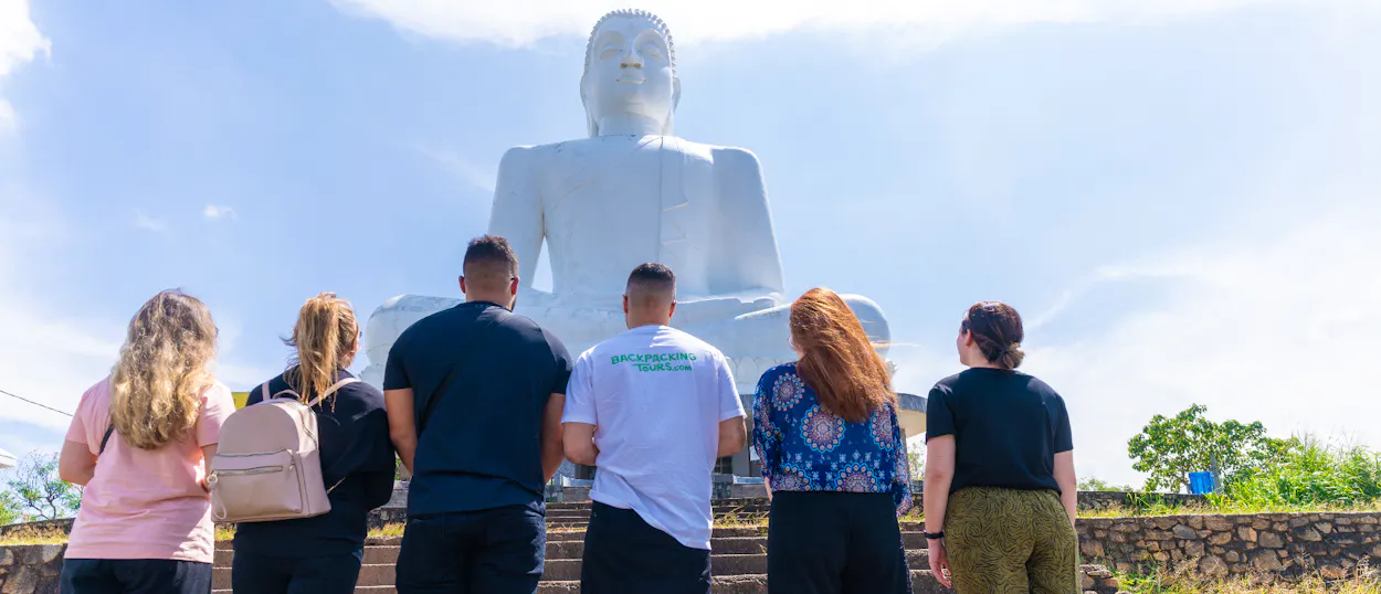 Six travelers look up at the massive white Buddha statue under a clear sky in Dambulla.