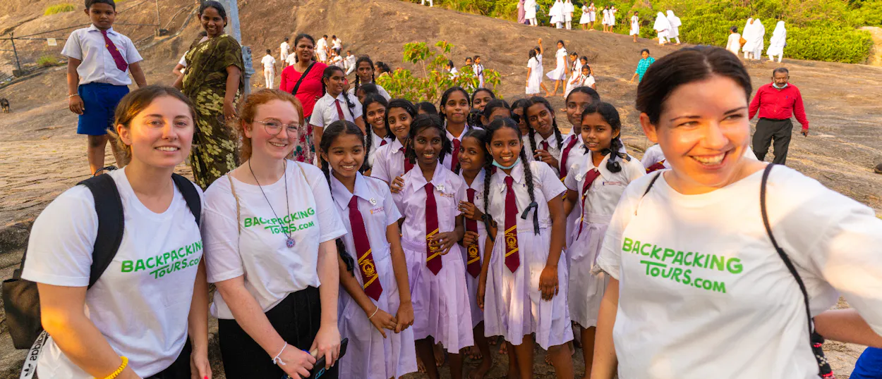 Three smiling backpackers pose with a group of happy Sri Lankan schoolgirls on a rocky hillside.