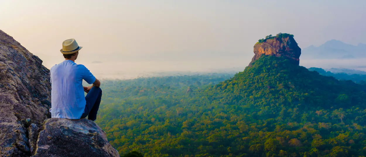 A person sits on a rocky outcrop, admiring the panoramic view of Sigiriya Rock and lush Sri Lankan forests.