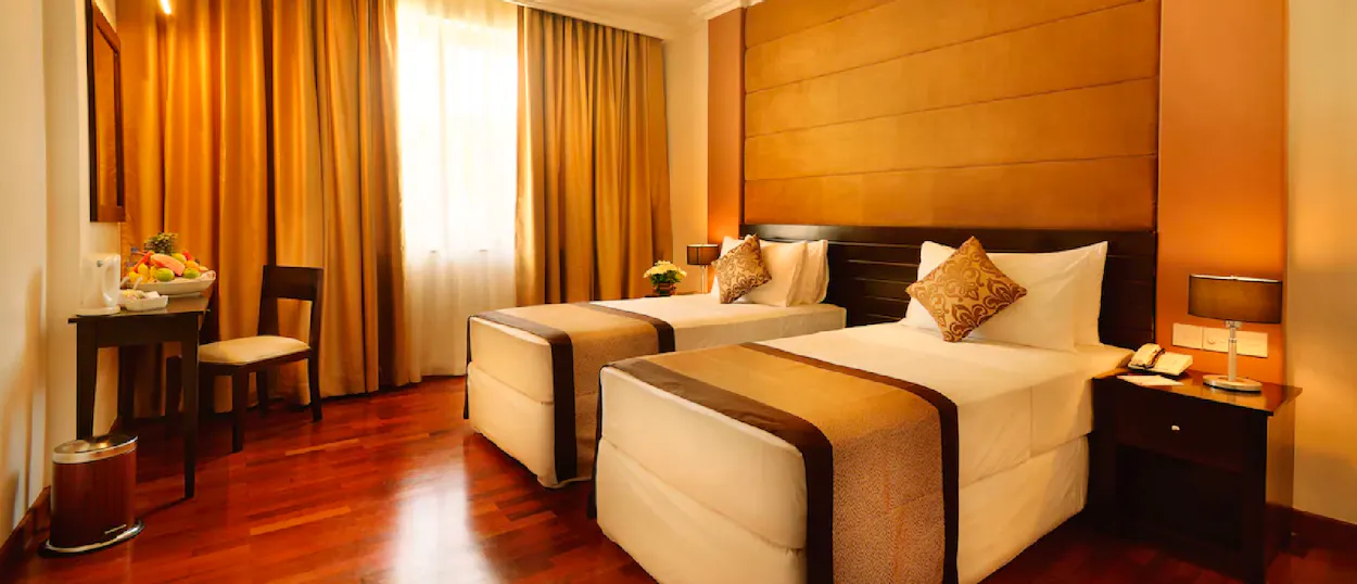 A comfortable hotel room with two single beds, wooden floor, and warm orange curtains.
