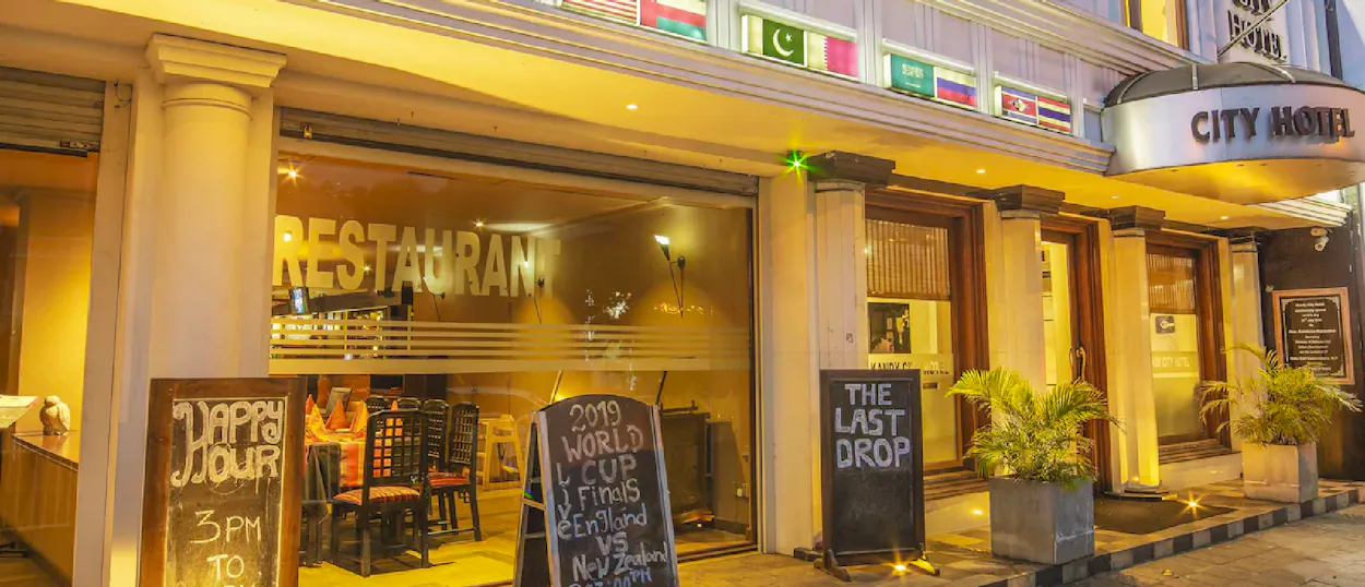 A brightly lit city hotel entrance with a restaurant, flags, and promotional chalkboards at night.