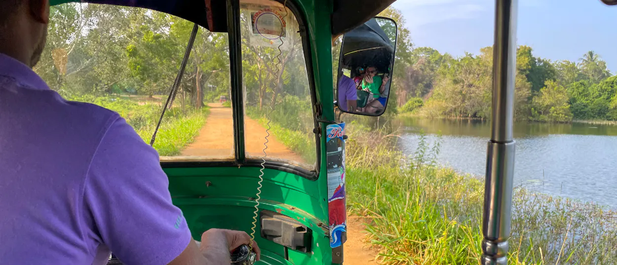 A person drives a green tuk-tuk along a dirt road next to a tranquil lake in rural Sigiriya.