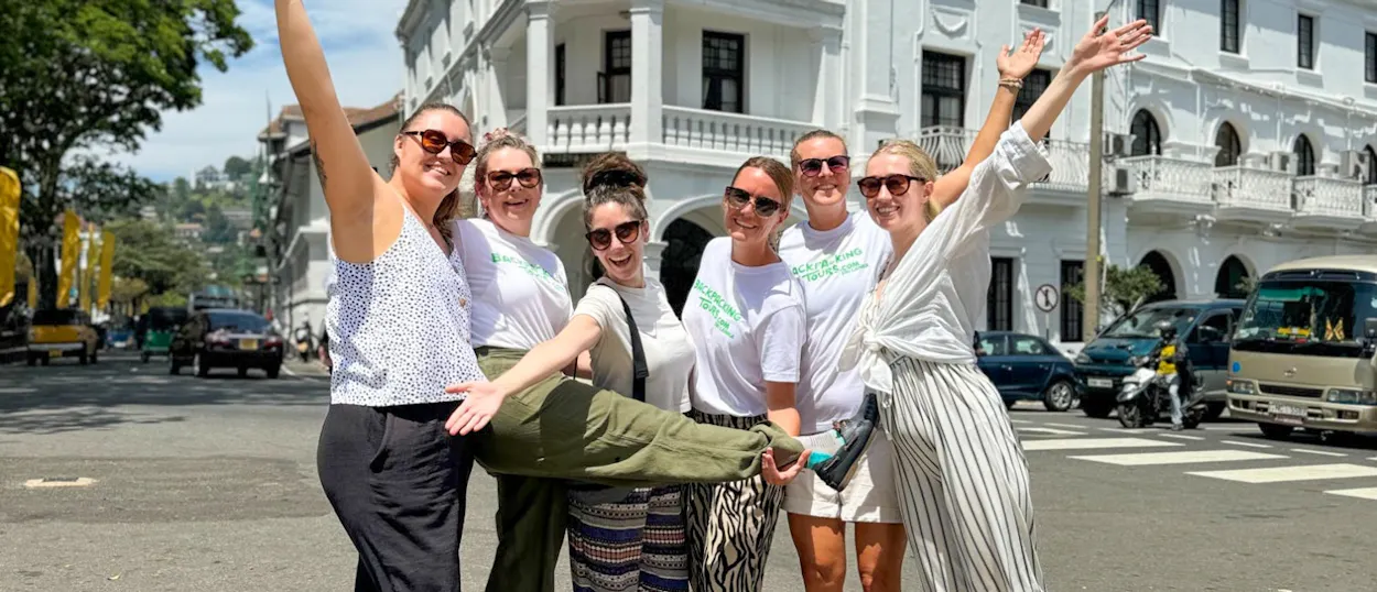 Six joyful friends pose on a sunny street in Kandy, Sri Lanka, playfully lifting one person.