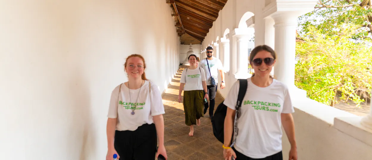 Four smiling backpackers walk along a bright white arched corridor with green trees in Kandy.
