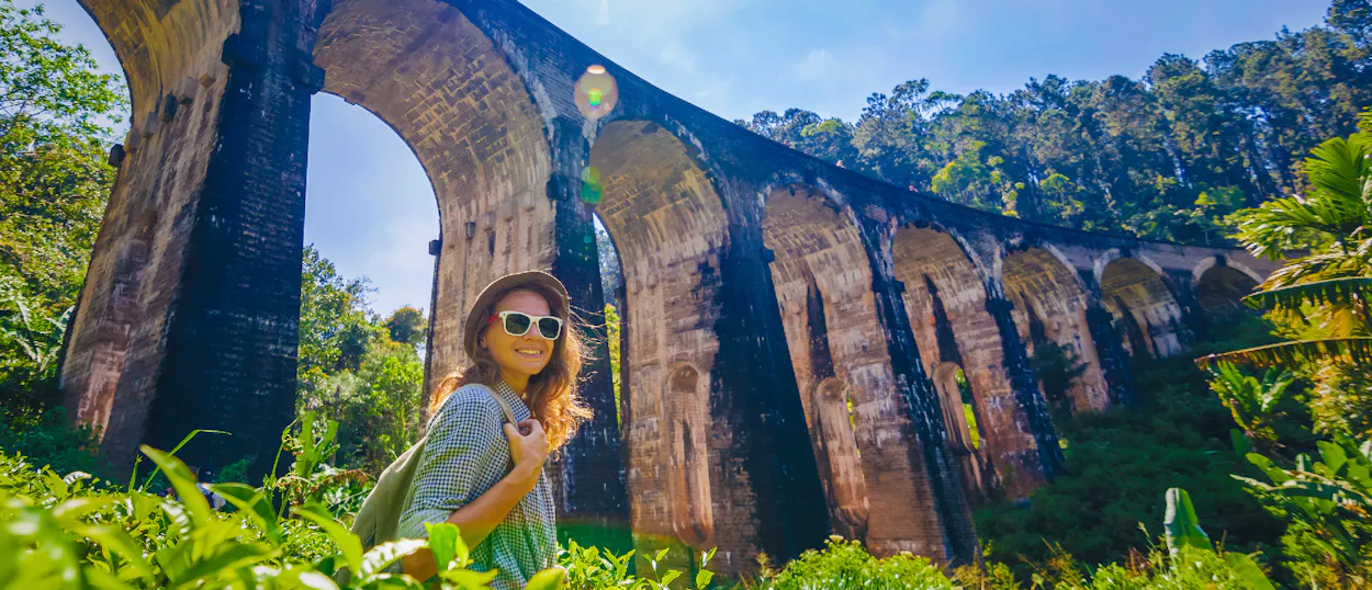 A smiling woman in sunglasses stands in green tea fields before the Nine Arch Bridge, Ella.