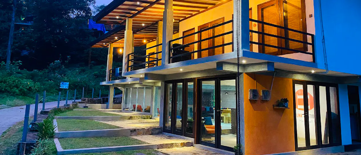 A modern guesthouse glows with warm lights at dusk, nestled in the lush Ella mountains.