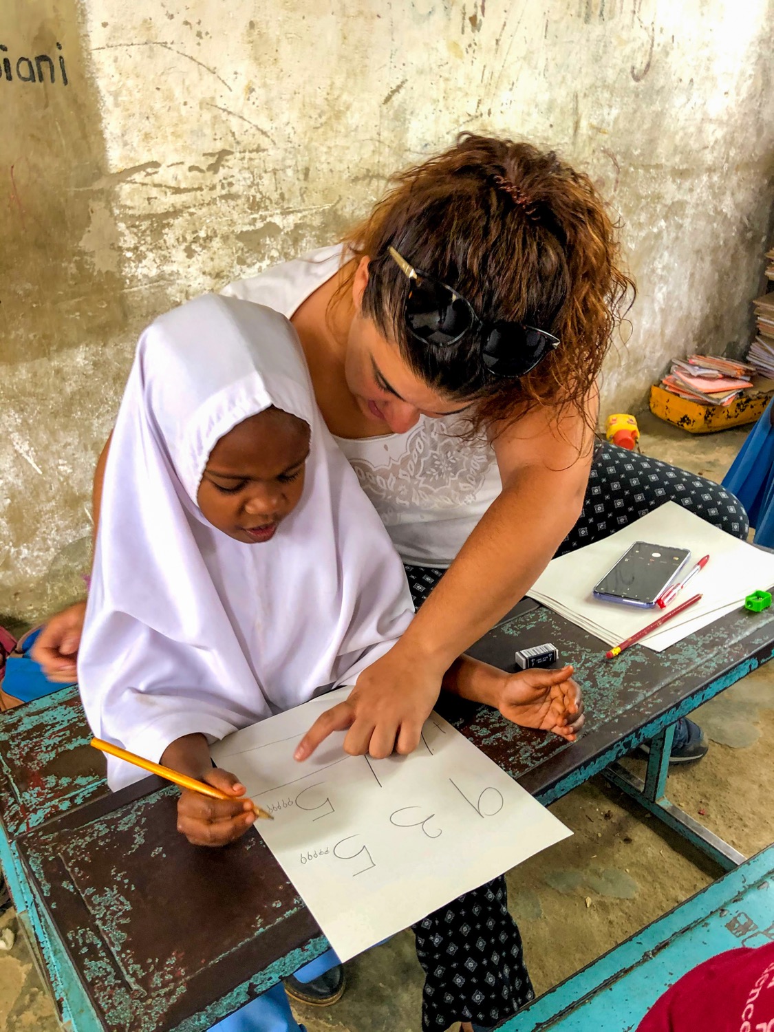 A volunteer teacher helps a young child practice writing numbers at a worn desk in Tanzania.