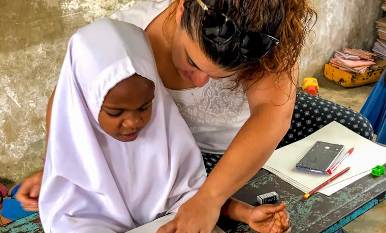 A volunteer teacher helps a young child practice writing numbers at a worn desk in Tanzania.