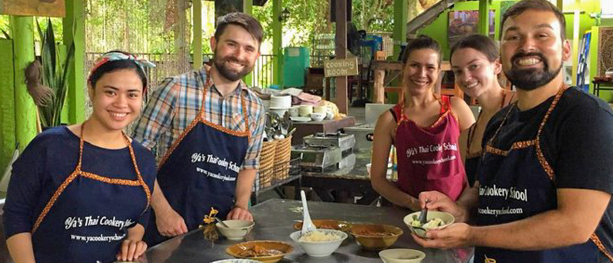 Five happy people in aprons learning to cook Thai food at a cooking school.