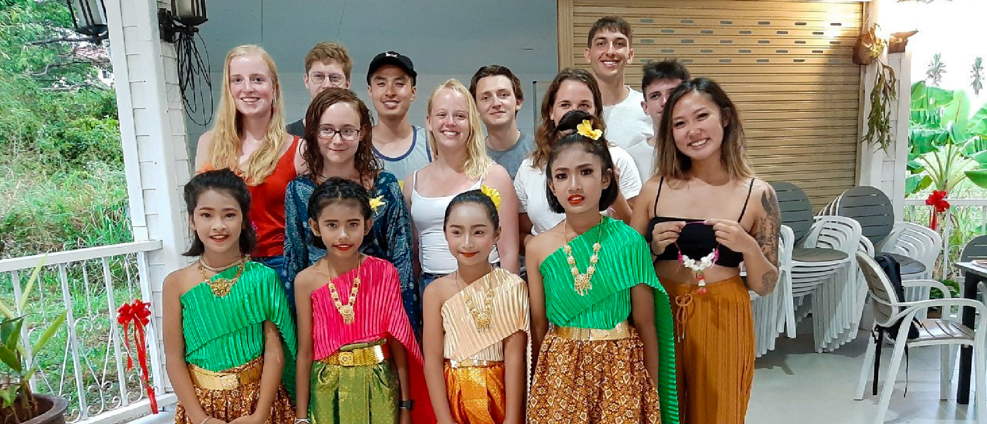 Smiling tourists and three young girls in traditional Thai dance costumes pose in Thailand.