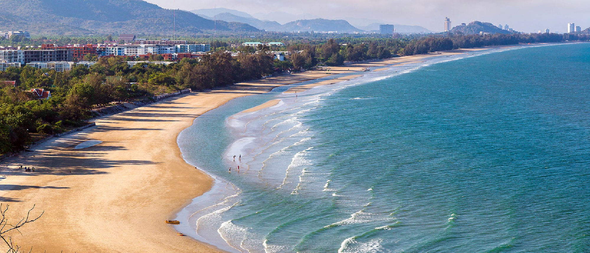 A long, curving sandy beach with turquoise waves, lush coastal trees, and distant buildings in Thailand.