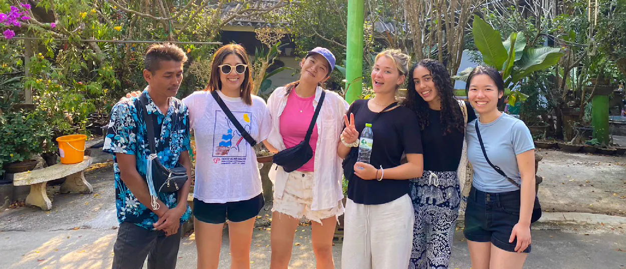 Six smiling people pose together outdoors in a sunny, green garden setting.