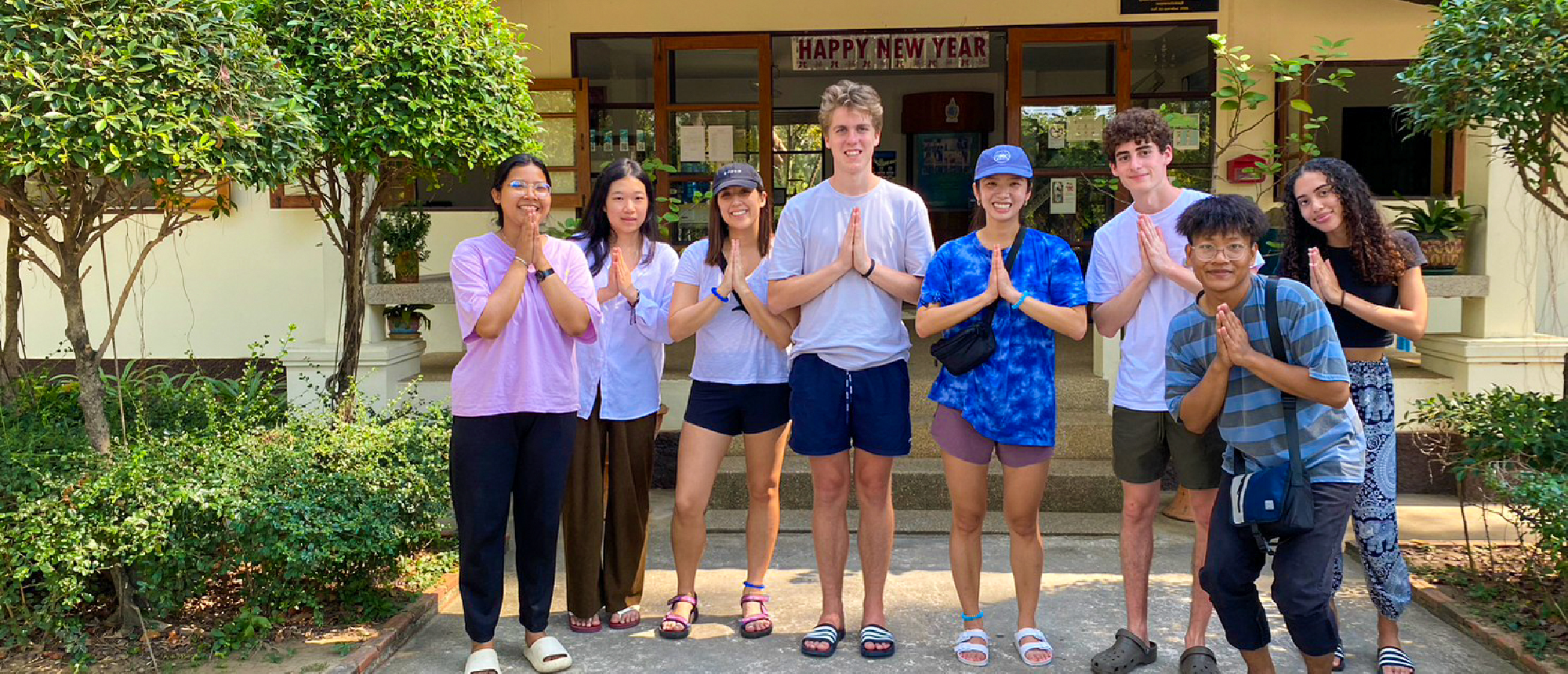 Eight smiling travellers perform the traditional Thai wai greeting outside a building in Thailand.