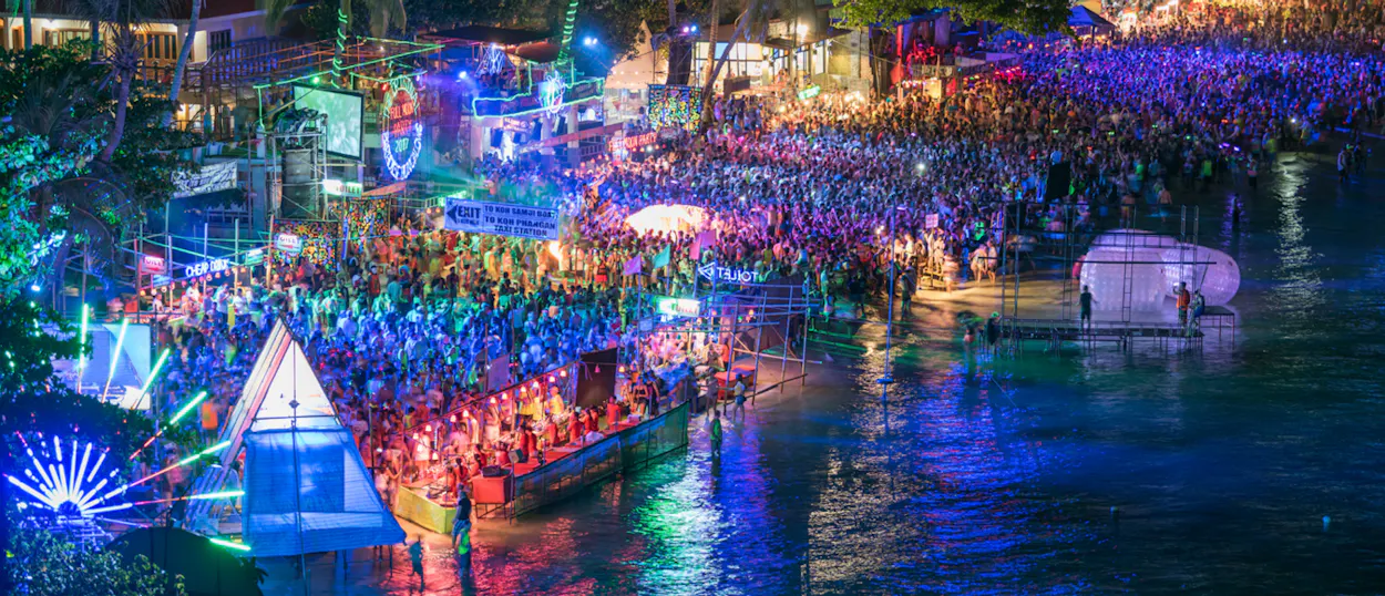 A massive full moon party on Koh Phangan beach, illuminated by vibrant neon lights and dancing crowds.