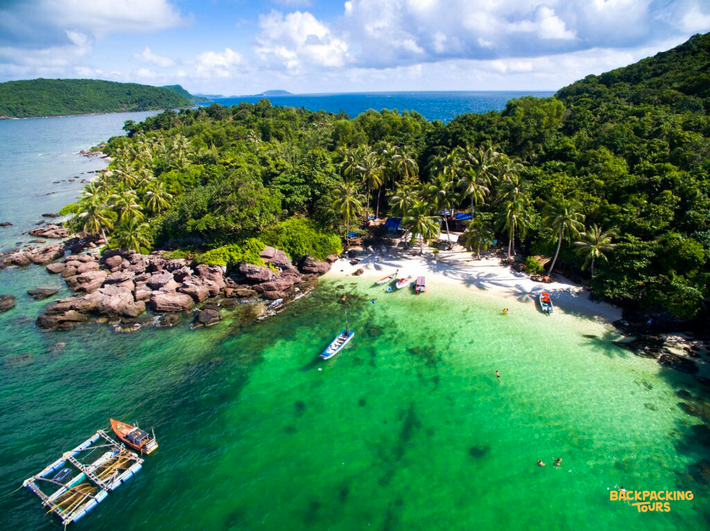 Boats float on clear emerald water beside a white sand beach and lush Koh Phangan island.