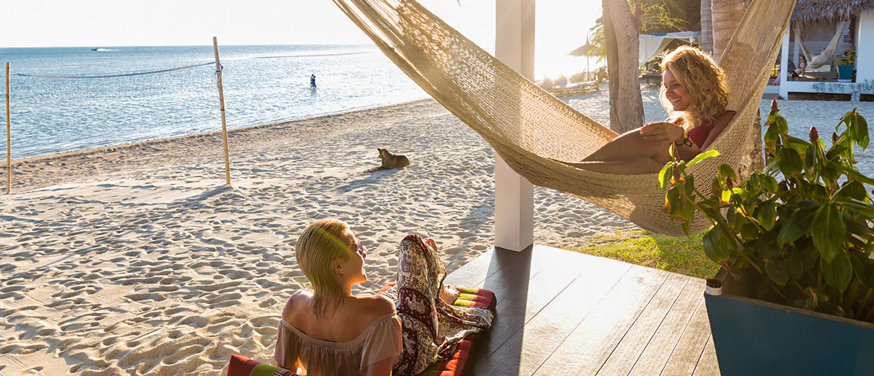 Two women relax on a porch overlooking a sunny beach and calm sea in Koh Phangan.
